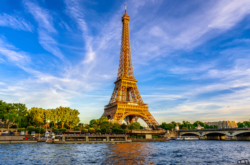 Image of the Eiffel tower with a river in the foreground under a bright blue sky.