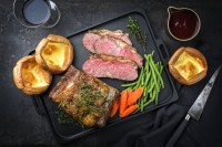 Image of sliced roast beef, popovers, carrots, and vegetables on a platter with a gravy boat against dark grey background.