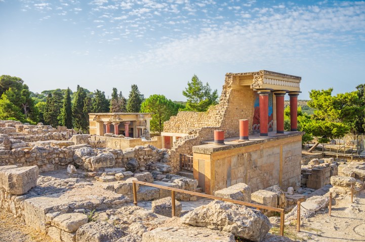 Image of stone ruins with red columns under light blue sky spotted with tiny white clouds.